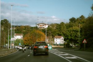 Vehicles navigate a rural road lined with scenic houses and lush trees under a clear sky.